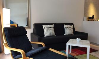 A cozy living room with a black sofa and cushions. There is a bright table and a chair on a colorful rug.