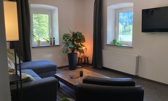 A cozy living room with a sofa, armchair, and a coffee table. The room has large windows with a view of greenery and a modern TV unit on the wall.