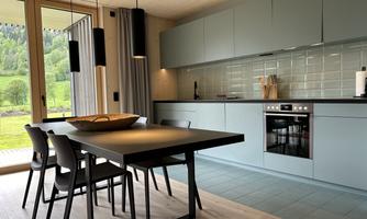 A modern kitchen with gray cabinets and a wooden dining table. Large windows provide a view of the green landscape.