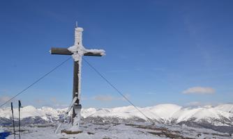 A wooden cross covered in snow stands on a mountain peak. In the background, snow-covered mountains and a clear blue sky are visible.