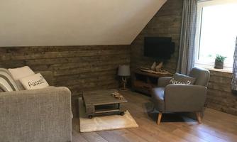 A cozy living room in the attic with wooden paneling. There is a sofa, an armchair, and a television, as well as large windows with a view outside.