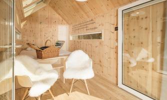 A bright, modern room design with wooden paneling. Two white chairs and a table are in the center, while a basket is lying on the bed.