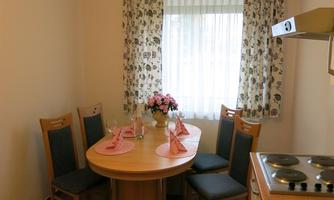 A cozy kitchen with a round dining table and chairs. The room has windows with curtains and a modern kitchenette.