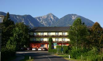 A pleasant hotel building with a green facade in front of impressive mountains. The surroundings are filled with trees and shrubs, exuding tranquility.