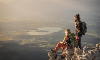 Two hikers are sitting on a rock, enjoying the view of a picturesque landscape. In the background, hills and a larger lake can be seen.