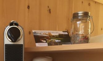 A modern coffee machine stands on a wooden table next to a glass with ingredients. In the background, there is a book and an information card.