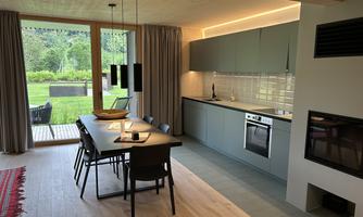 Modern kitchen with a dining area and large windows overlooking a green garden. The space is bright and inviting, with stylish furnishings.