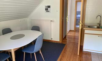 A cozy dining area with a round table and blue chairs. In the background, a kitchenette and an open bedroom are visible.