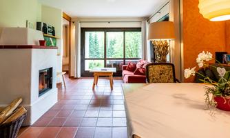 A bright living room with large windows and a view of the garden. There is a cozy couch, a table, and a fireplace.