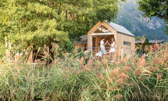 A charming wooden house amidst lush plants and trees. Two people are standing on the porch and enjoying nature.