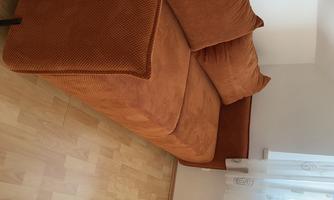 A cozy orange couch with large pillows is positioned in a bright room. It is placed next to a window and has a light wooden floor.