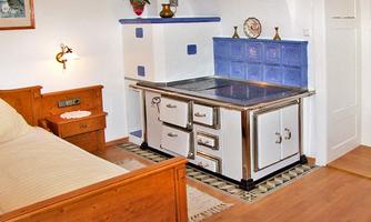A cozy room with a wooden bed and a historic stove. The walls are painted in light colors, and there are decorative elements like plants and pictures.