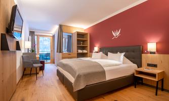A modern hotel room with a large bed, cozy chairs, and a balcony. The walls are painted in warm colors, and there are wooden surfaces for an inviting atmosphere.
