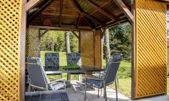 A cozy pavilion with a table and chairs, surrounded by beautiful greenery. The walls are partially clad with wooden planks.