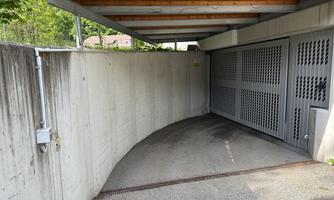 A curved driveway to a garage with grey walls and a lattice privacy screen. Covered by a wood shed, surrounded by greenery.