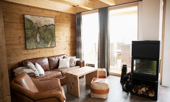 A modern living room with a leather couch, a wooden table, and a fireplace. Large windows provide plenty of light and a view outside.