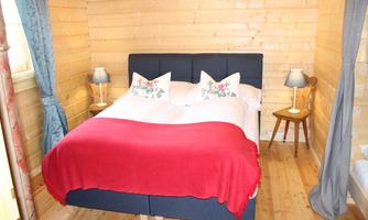 A cozy bedroom with a double bed and a red bedspread. The walls are made of wood, and there are two nightstands with lamps.