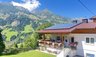 A cozy house with a red roof and solar panels, surrounded by green mountains. The sky is blue and the landscape is idyllic.