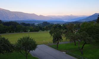 A quiet landscape farm with green meadows and trees. In the background, there are gentle mountains and a clear sky.