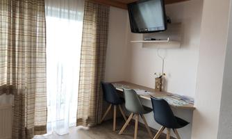 A cozy room with a table and chairs. In the background, there are large windows with curtains and a television on the wall.