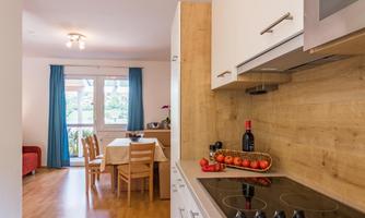 A modern kitchen with wooden elements and a dining table. In the background, there are windows with curtains and a cozy seating area.