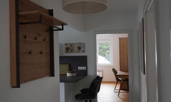 A bright hallway with a modern lampshade and a wooden shelf. In the background, another room with a dining table is visible.