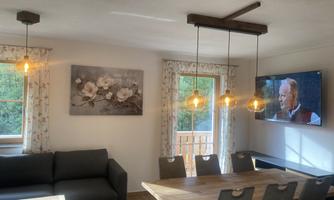 A cozy living space with a large dining table and a couch. A television hangs on the wall, and the windows let in plenty of daylight.