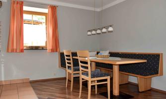 A cozy dining area with a wooden table, surrounded by bench-like seating and chairs. Light curtains frame the window and let in plenty of light.