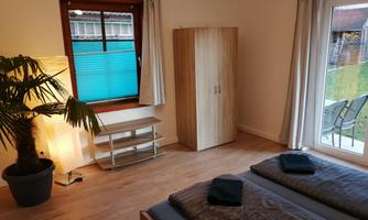 A bright bedroom with two beds and a wooden wardrobe. A window overlooking the garden and a plant create a cozy atmosphere.