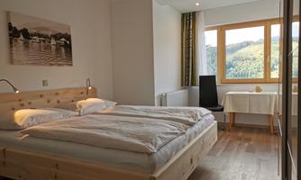 A cozy bedroom with a large wooden bed and two pillows. The room has large windows overlooking a green landscape.