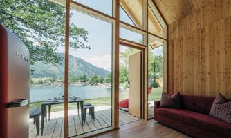 A modern wooden house with large windows and a view of a lake. Outside, tables and chairs are placed on a wooden terrace, surrounded by green nature.