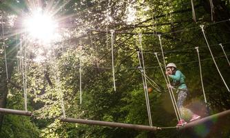 A girl is climbing on a high ropes course between trees. The sun is shining through the foliage, creating a bright, friendly atmosphere.