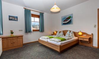 A cozy bedroom with a double bed and wooden furniture. The windows are adorned with blue curtains, and a beautiful boat print hangs on the wall.