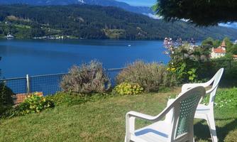 A beautiful view of a lake with mountains in the background. Two white plastic chairs are positioned on the grass.