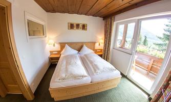 A cozy bedroom with a wooden double bed and beautiful pillows. The room has large windows that lead to a balcony overlooking nature.