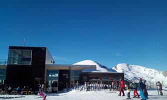 A ski resort with a modern building in the foreground. In the background, there are snow-covered mountains and people skiing.