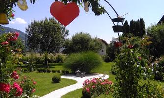 A beautiful garden with colorful roses and a small path. A red heart hangs above the garden and it radiates sunshine.
