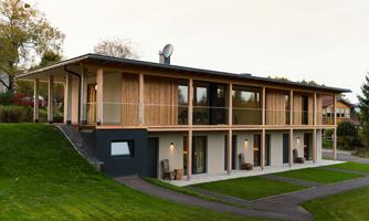 A modern house with wooden cladding and large windows. The surroundings are green and inviting, ideal for relaxation.