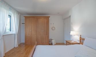 A bright bedroom with a large bed and a wooden wardrobe. The walls are white and there are curtains that let in natural light.