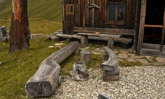 A rustic wooden cabin with benches made from tree trunks and a small table. The surroundings are designed with grass and stones.