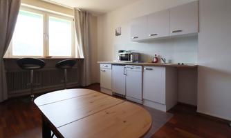 A bright kitchen unit with modern furniture and a wooden dining table. Large windows provide plenty of daylight.