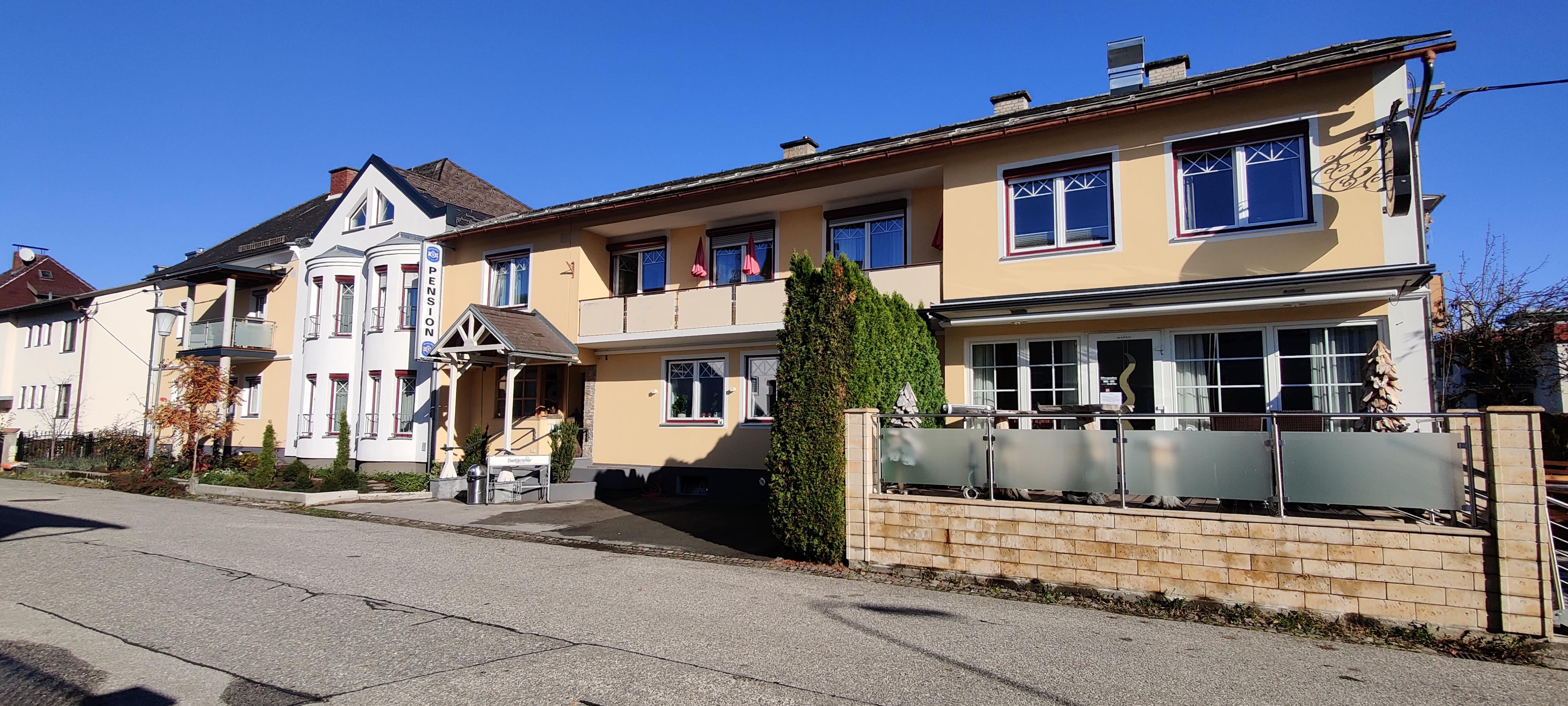 A charming building with a yellow facade and large windows. In front of the house, there are cozy seating areas and a well-maintained garden.