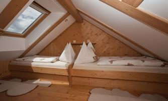 A cozy attic room with wooden paneling and two beds. The window lets in daylight and offers a view of the sky.