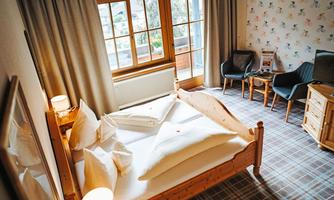 A cozy bedroom with a large bed and bright curtains. The room is furnished with two chairs and a table and has windows overlooking the outside.