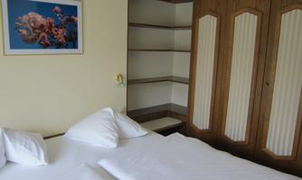 A simple bedroom with two beds and white bedspreads. In the background, there is a wardrobe and a picture of flowers on the wall.