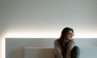 A thoughtful woman sits on a bright bench in a minimalist room. Soft light emphasizes the calm atmosphere.