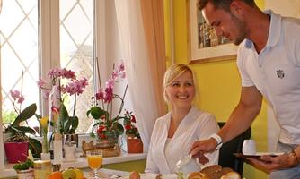 A friendly breakfast in a bright room with plants. A man serves a smiling woman a meal at the table.