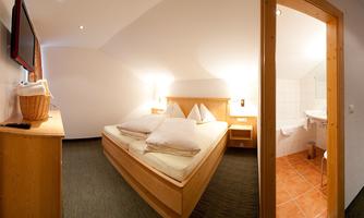 A cozy hotel room with a double bed, wooden furniture, and a television. To the right, the bathroom is visible, which is modernly furnished.