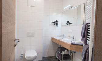 A modern bathroom with white tiles and a wooden sink cabinet. It includes a toilet, a mirror, and towels.