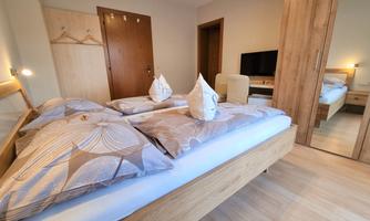 A modern room with a double bed and beige walls. There is a television and a large mirror that reflects the space.